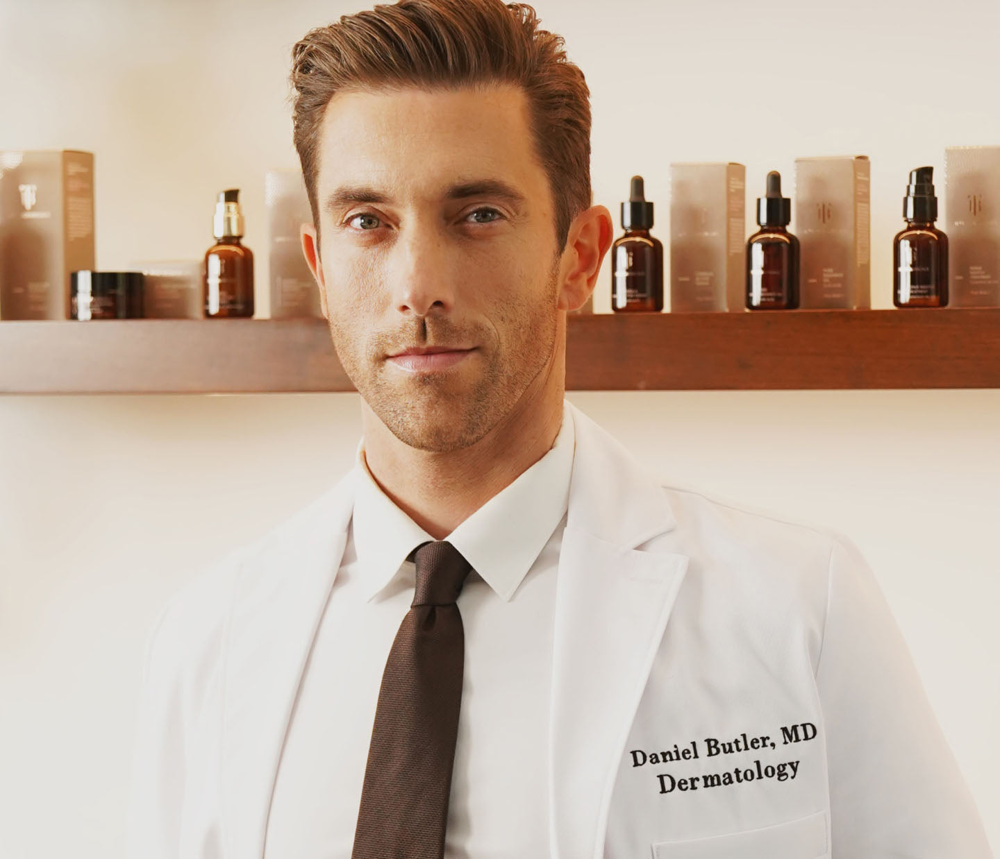 A man wearing a white lab coat embroidered with "Daniel Butler, MD, Dermatology" stands confidently in front of a wooden shelf. The shelf is lined with amber glass bottles and boxes of True Botanicals skincare products. He has neatly styled hair and a professional demeanor, with a neutral-toned background that emphasizes a clean, clinical setting.